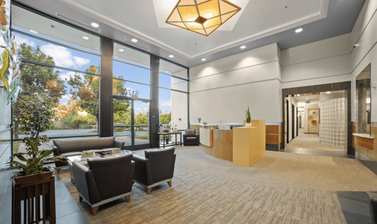 Modern office lobby with seating area, reception desk, large windows, and indoor plants.